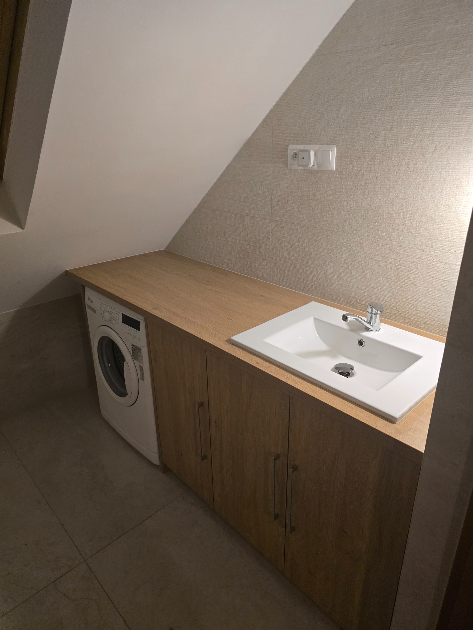 Zabudowa pralki i umywalki w łazience na poddaszu. Blat z drewnopodobnego laminatu, szafki z frontami w kolorze jasnego drewna. Umywalka ceramiczna, bateria chromowana.