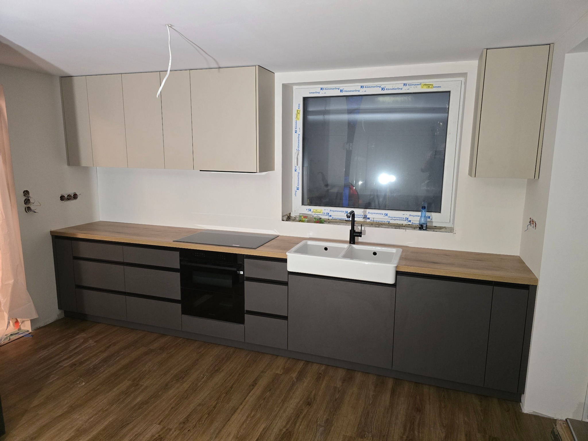 Nowoczesna kuchnia: szare szafki dolne z drewnianym blatem, biała umywalka farmerska, beżowe szafki górne. Widoczne gniazdka elektryczne i niedokończone wykończenie.