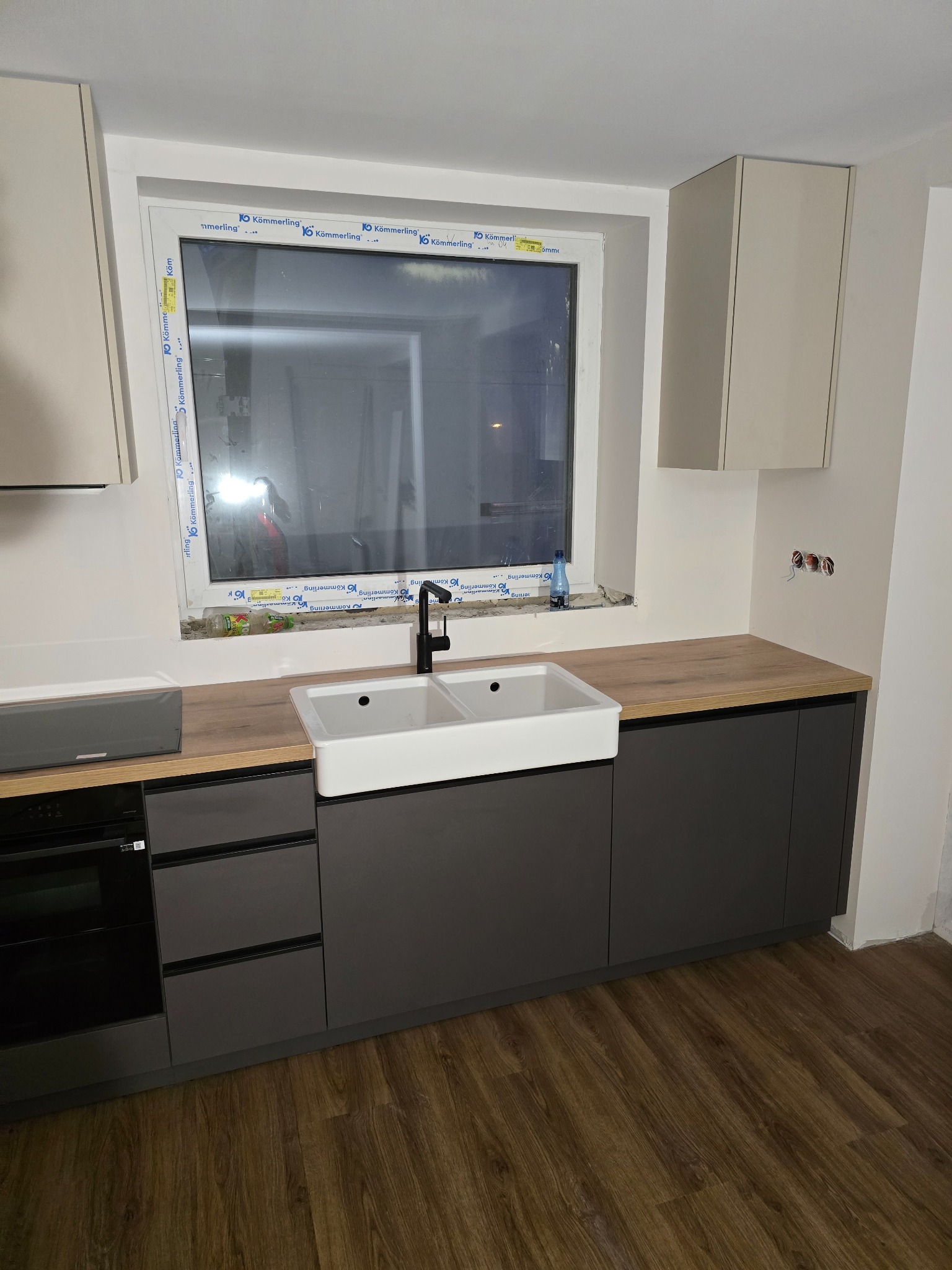 Szara zabudowa kuchenna z drewnianym blatem i białym zlewem farmerskim pod oknem z ramą Kömmerling. Widoczne szafki wiszące i sprzęt AGD.