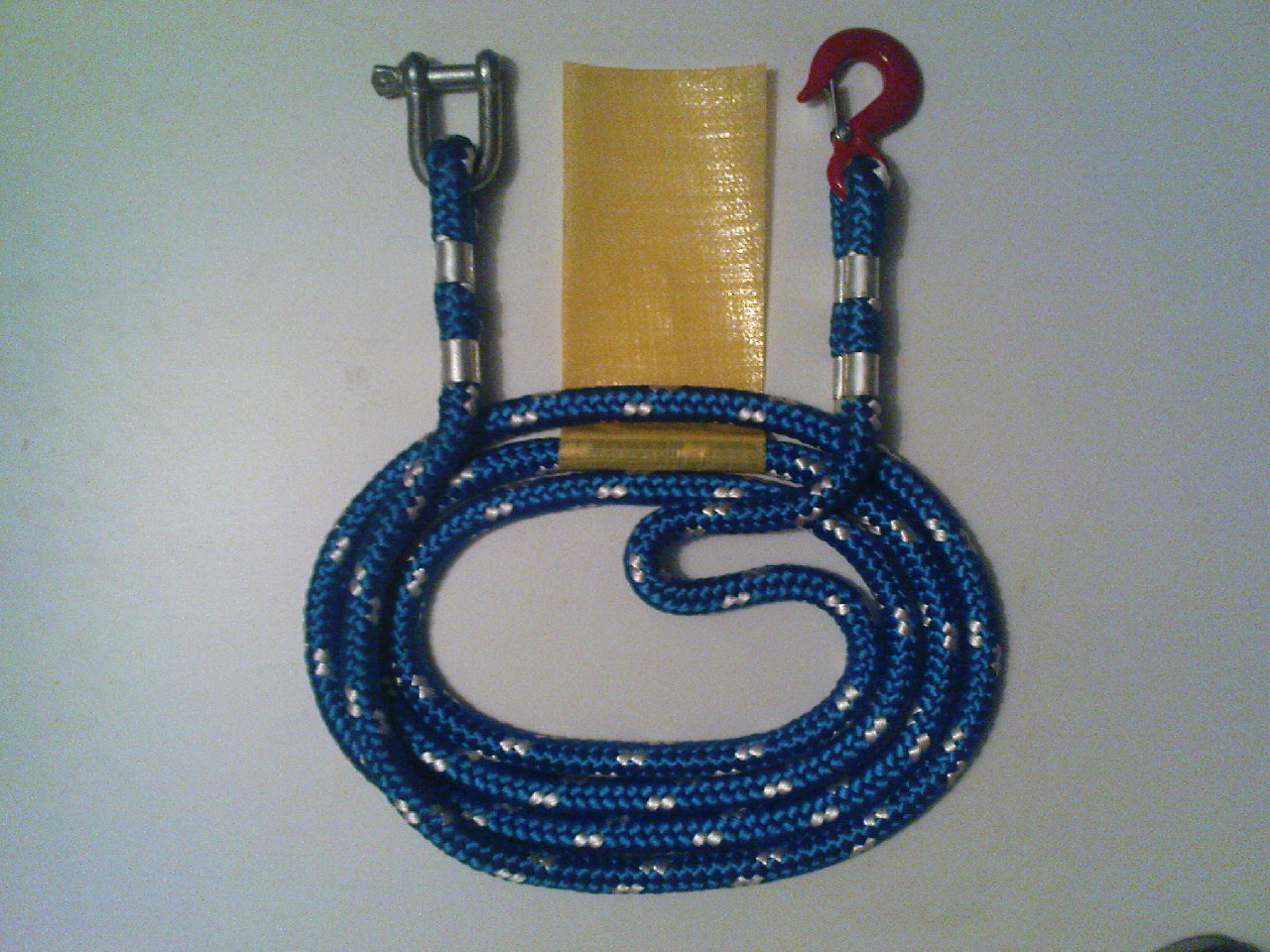 Niebieska lina holownicza z metalowym szeklem z jednej strony i czerwonym hakiem z drugiej, zwinięta w owal na białym tle, obok żółty prostokątny element.