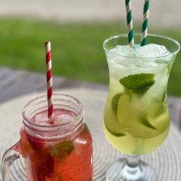 KOZACKIE JADŁO OKSANA MAKSYMENKOVA - Dwa drinki: truskawkowe mojito w słoiku z uchwytem i klasyczne w kieliszku, oba z lodem, miętą i słomkami na plecionej podkładce. Tło rozmyte, zielone.