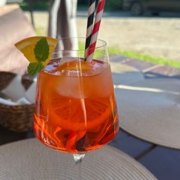 KOZACKIE JADŁO OKSANA MAKSYMENKOVA - Aperol Spritz w kieliszku z plasterkiem pomarańczy i miętą, dwie słomki w paski, na beżowej podkładce. W tle zieleń i samochód.