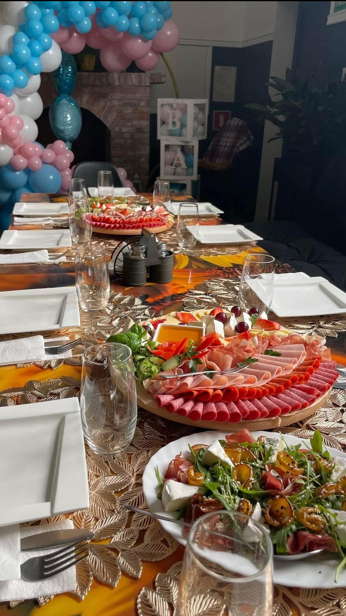 Stół z dekoracją balonową i aranżacją przekąsek: wędliny, sery i sałatki na drewnianych deskach. Nakrycia i szkło przygotowane do przyjęcia.