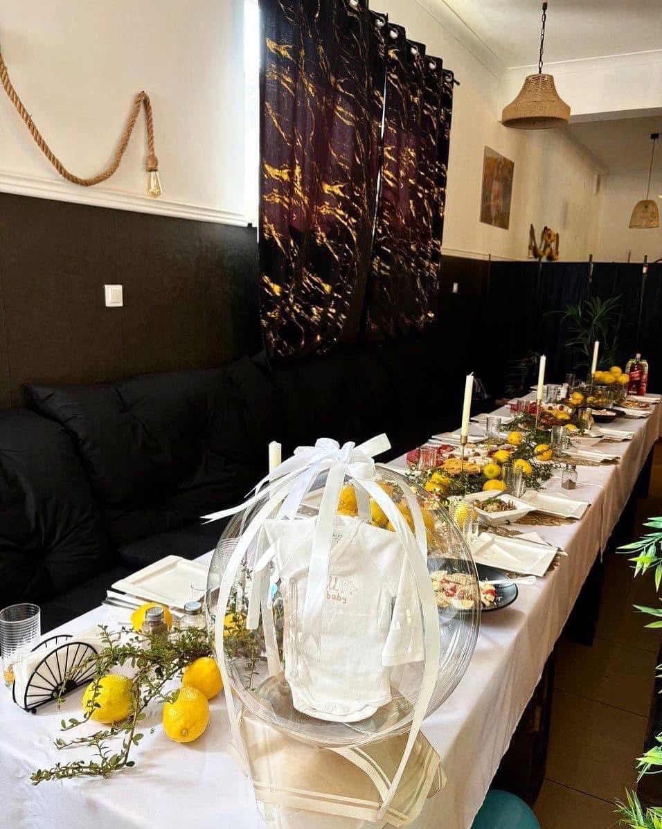 Stół udekorowany na baby shower z białym body w przezroczystym balonie, cytrynami i świecami. Czarny narożnik, ciemne zasłony i dekoracja ze sznura w tle.