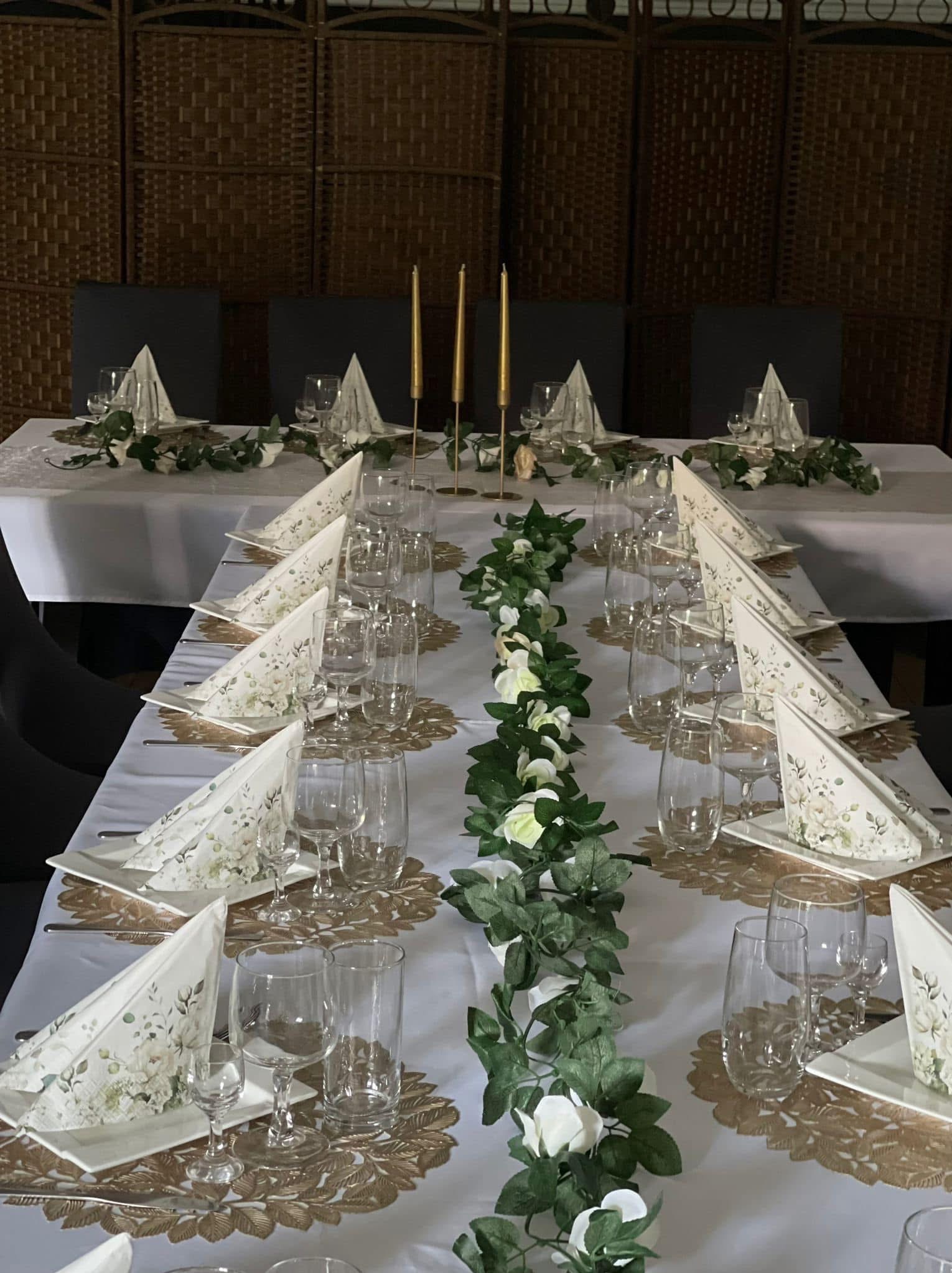 Elegancko nakryty stół z białym obrusem, złotymi podkładkami, kwiatową girlandą i serwetkami w kwiaty, gotowy na przyjęcie. W tle parawan.