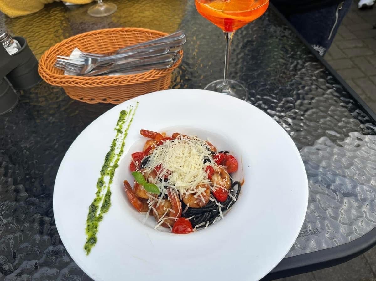 Elegancko podane czarne spaghetti z krewetkami, pomidorami i serem na białym talerzu. W tle napój pomarańczowy i koszyk ze sztućcami.