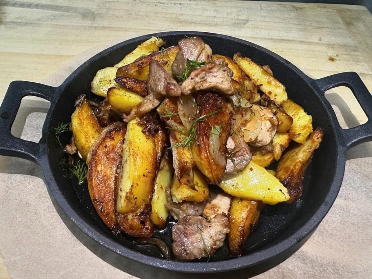 Aromatyczne ziemniaki pieczone z mięsem i ziołami w czarnej patelni, gotowe do podania. Apetyczny widok dania na drewnianym stole.