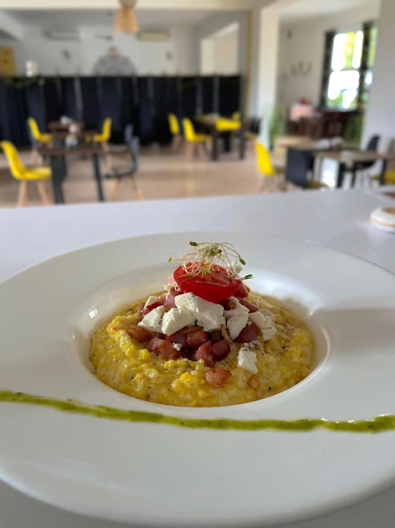 Elegancko podana potrawa z polenty, boczku, sera feta i pomidora, udekorowana kiełkami, na białym talerzu z zielonym sosem, w restauracyjnym wnętrzu.