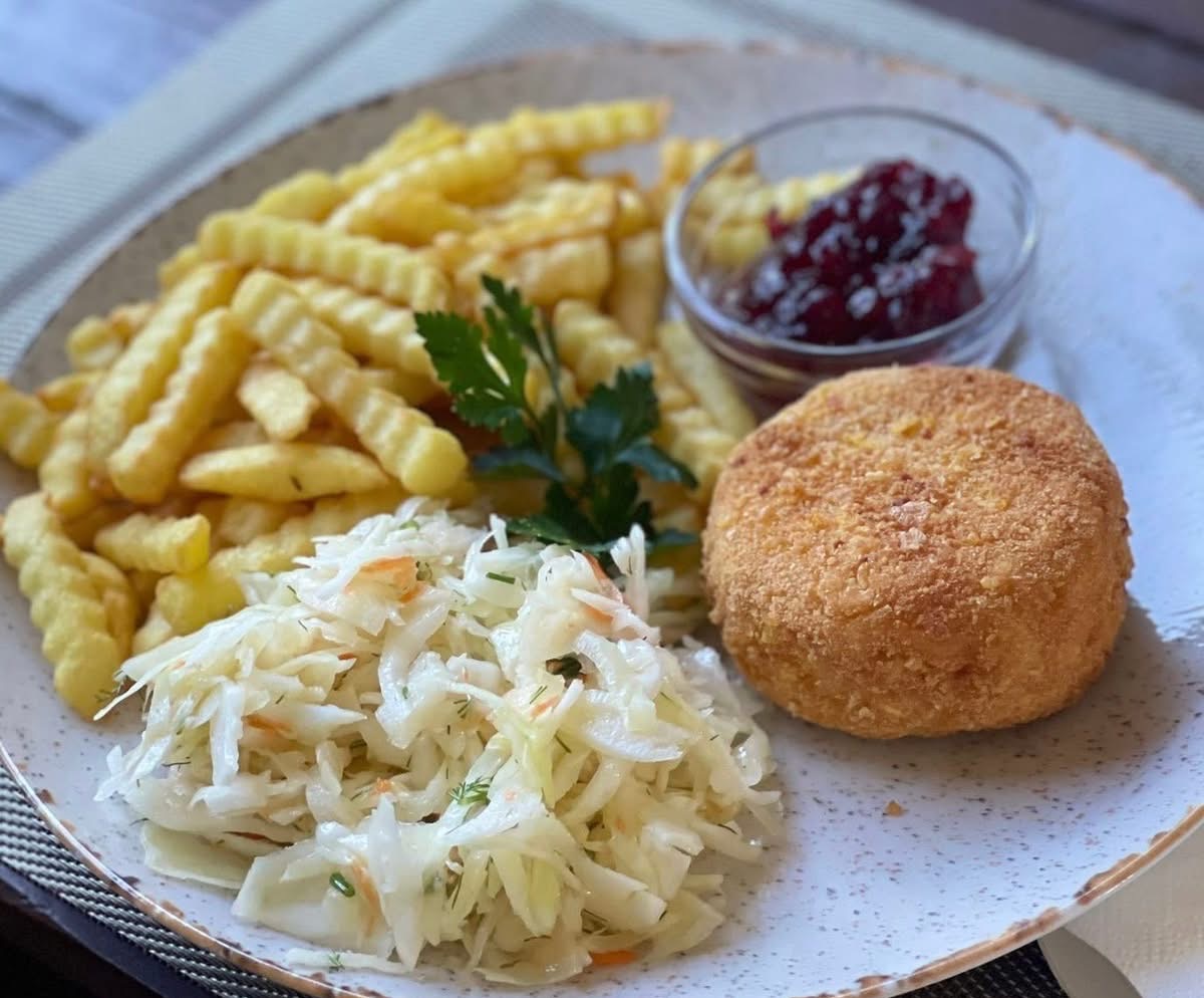 Zestaw obiadowy na talerzu: kotlet panierowany, frytki karbowane, surówka z białej kapusty i sos żurawinowy w szklanej miseczce, udekorowany świeżą natką pietruszki.