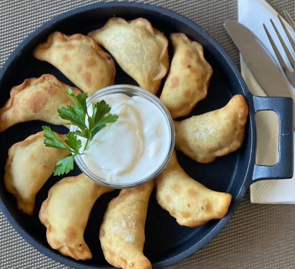 Ręcznie lepione pierogi z farszem, ułożone wokół miseczki ze śmietaną, udekorowane zieloną pietruszką na czarnym talerzu. Obok widoczne sztućce.