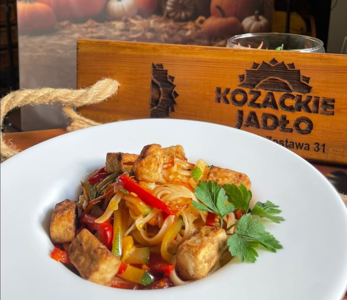 Azjatyckie danie z tofu i warzywami na białym talerzu, z drewnianym pudełkiem 'Kozackie Jadło' w tle i dekoracją z dyni. Apetyczne i kolorowe.