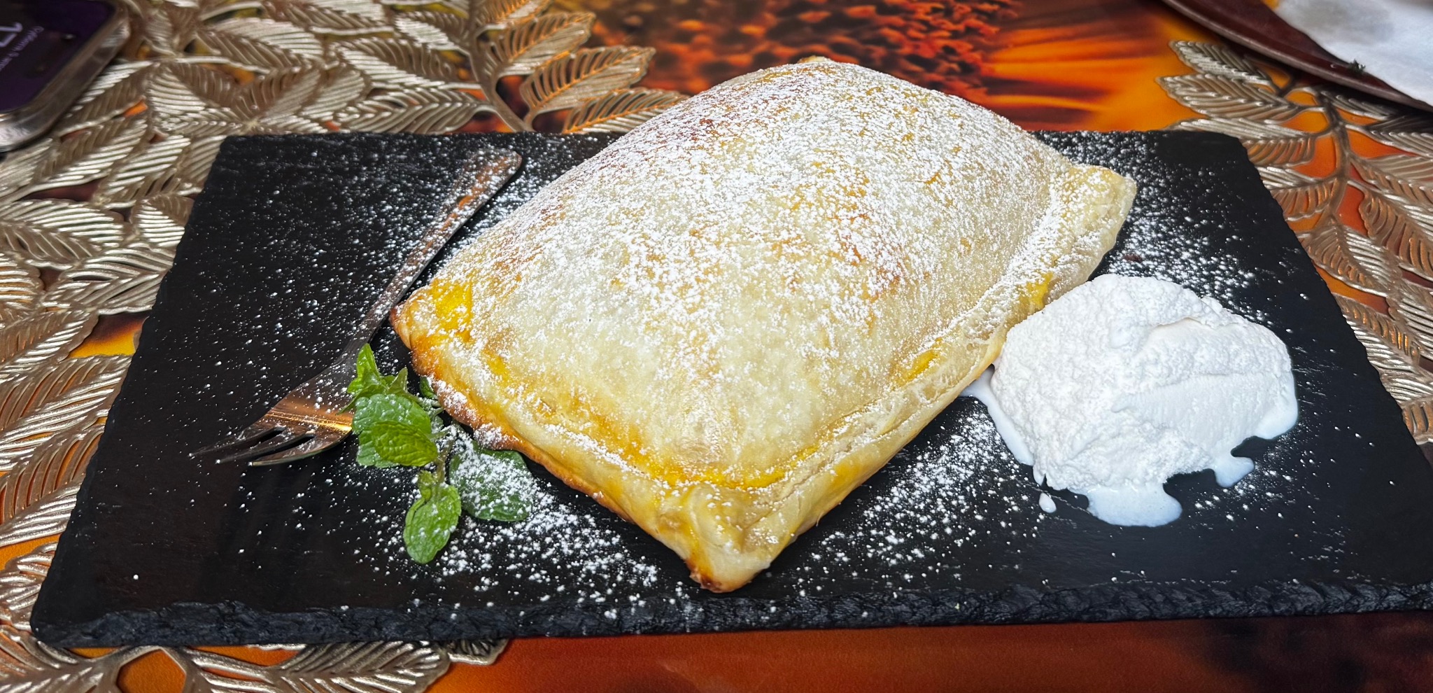 Apetyczny strudel jabłkowy posypany cukrem pudrem, podany z bitą śmietaną i miętą na czarnym talerzu, elegancko udekorowany. Idealny deser dla firm.