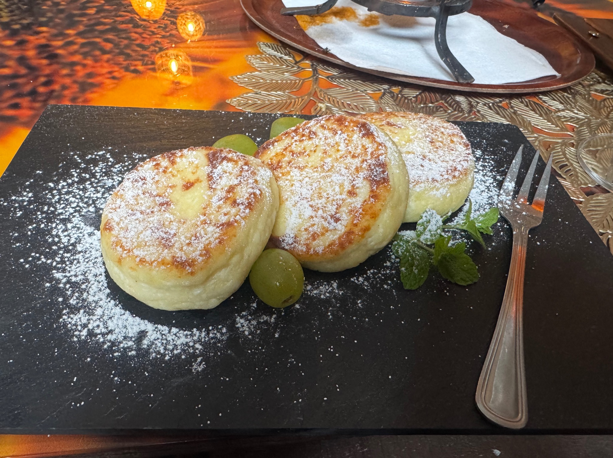 Trzy serniczki posypane cukrem pudrem na czarnym talerzu, ozdobione winogronami i miętą, obok widelca. Dekoracyjny stół w ciepłych barwach w tle.