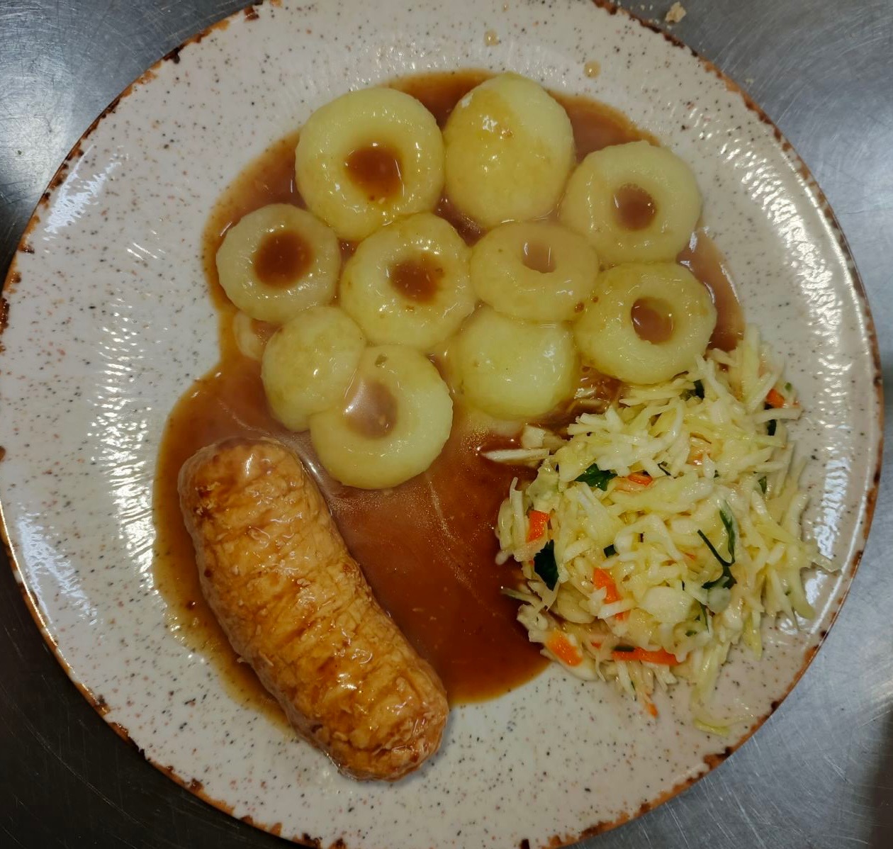 Kluski śląskie z sosem, pieczoną kiełbasą i surówką na talerzu z brązowym obramowaniem, ujęcie z góry, oświetlenie dzienne.