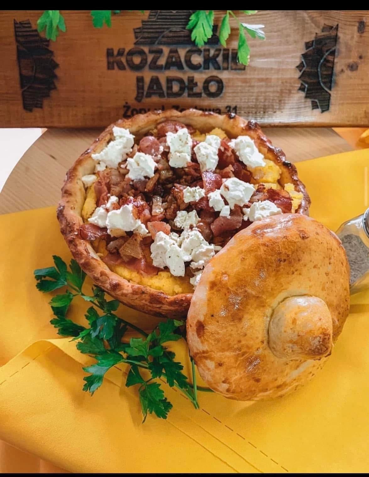 Kozacki placek z boczkiem i serem, podany z bułeczką na żółtej serwetce, dekorowany zieloną pietruszką. W tle drewniany szyld z nazwą lokalu.