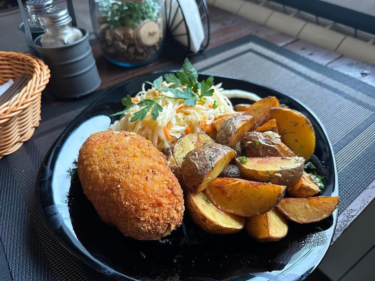 Czarny talerz z krokietem, ziemniakami i surówką, na tle szklanych pojemników i koszyka. Ujęcie z góry, światło dzienne.
