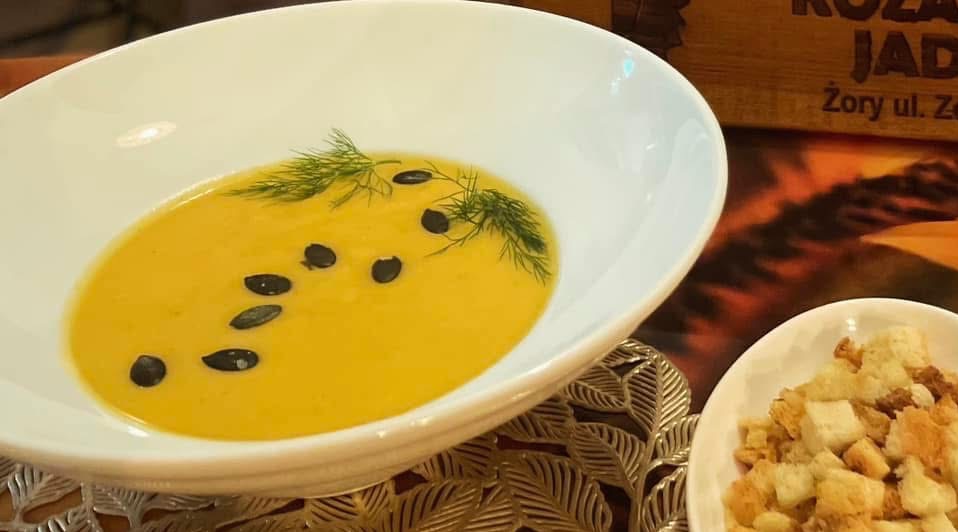 Krem z dyni w białej misce, dekorowany koperkiem i pestkami dyni, obok grzanki. W tle logo restauracji.