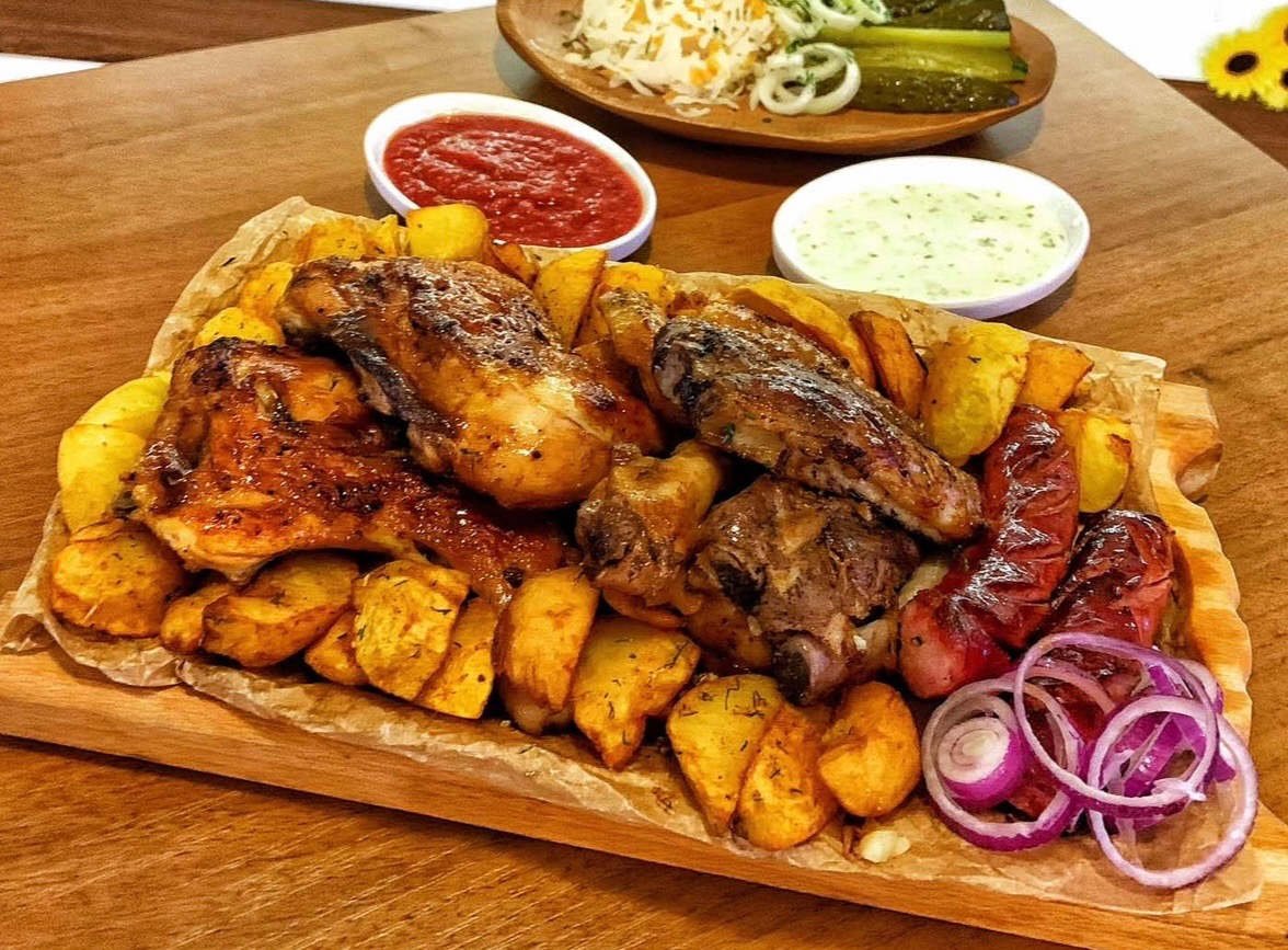 Bogato zastawiona drewniana deska: pieczone mięsa, ziemniaki, kiełbasa, czerwona cebula, sosy i sałatka na tle drewnianego stołu. Ujęcie z góry.