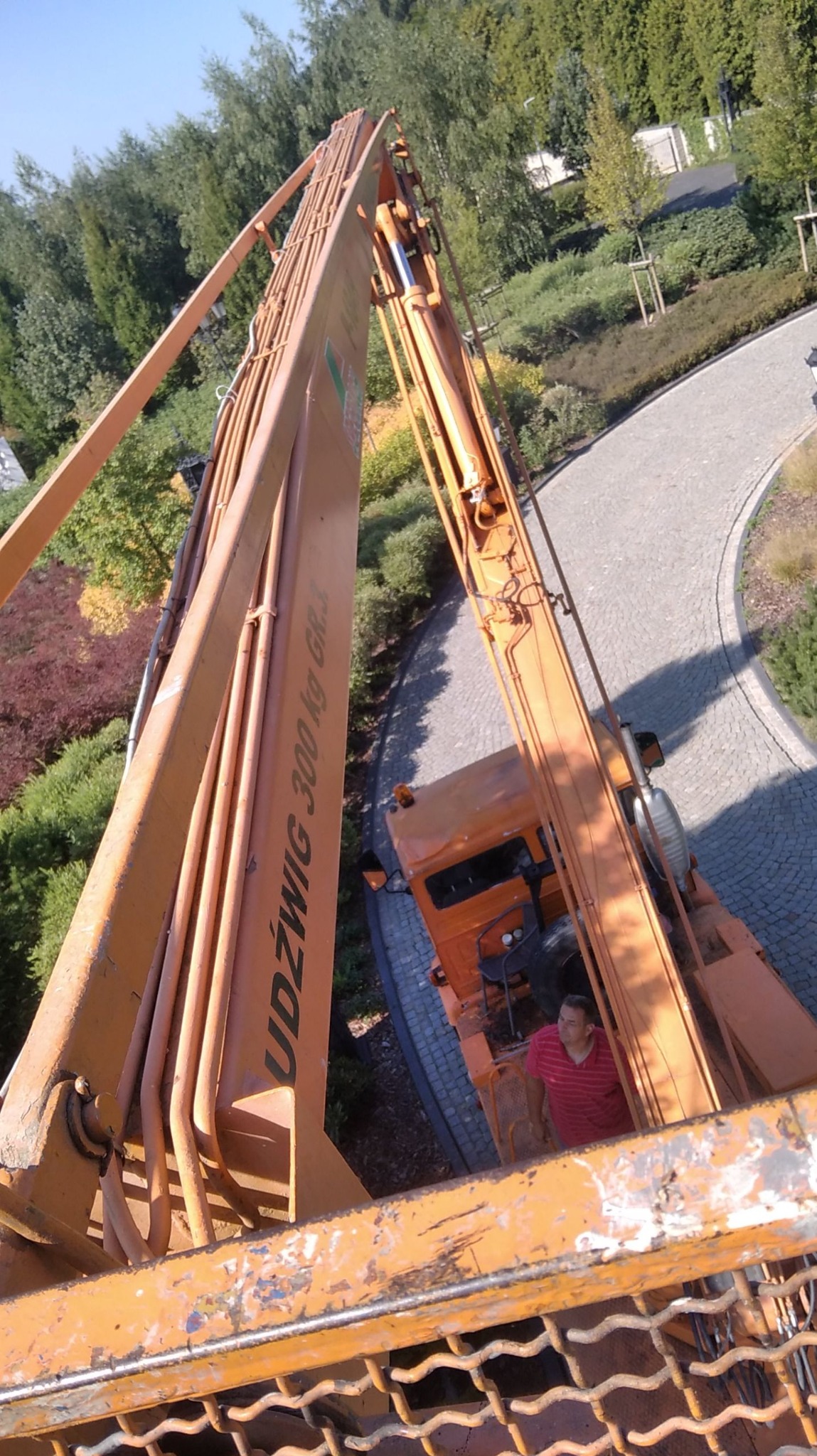Pomarańczowy podnośnik koszowy z operatorem na tle zieleni i brukowanej ścieżki, widok z góry z kosza. Maszyna marki Udźwig 300 kg.