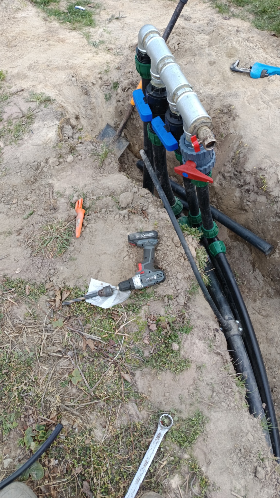 Instalacja systemu nawadniania w ziemi: czarne rury, metalowe złączki, zawory z niebieskimi i czerwonymi uchwytami. Obok narzędzia: wiertarka, klucze, łopata.