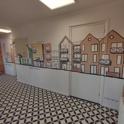 EFEKT - Wnętrze z dekoracyjnym malunkiem na ścianie przedstawiającym budynki w stylu szkicowym, uzupełnione napisem. Podłoga wyłożona wzorzystymi płytkami.