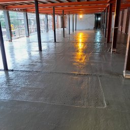 EFEKT - Świeżo wylana, mokra posadzka betonowa w surowym wnętrzu z metalową konstrukcją stropu i kolumnami. Widoczne refleksy światła na gładkiej powierzchni betonu.