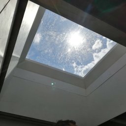 EFEKT - Wnętrze z oknem dachowym, widok na błękitne niebo z chmurami. Białe ściany i rama okna, widoczne ślady montażu. Ujęcie z dołu, fragment głowy osoby w kadrze.