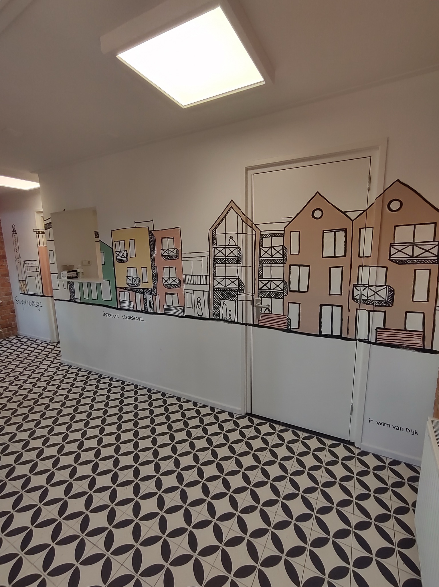 Wnętrze z dekoracyjnym malunkiem na ścianie przedstawiającym budynki w stylu szkicowym, uzupełnione napisem. Podłoga wyłożona wzorzystymi płytkami.