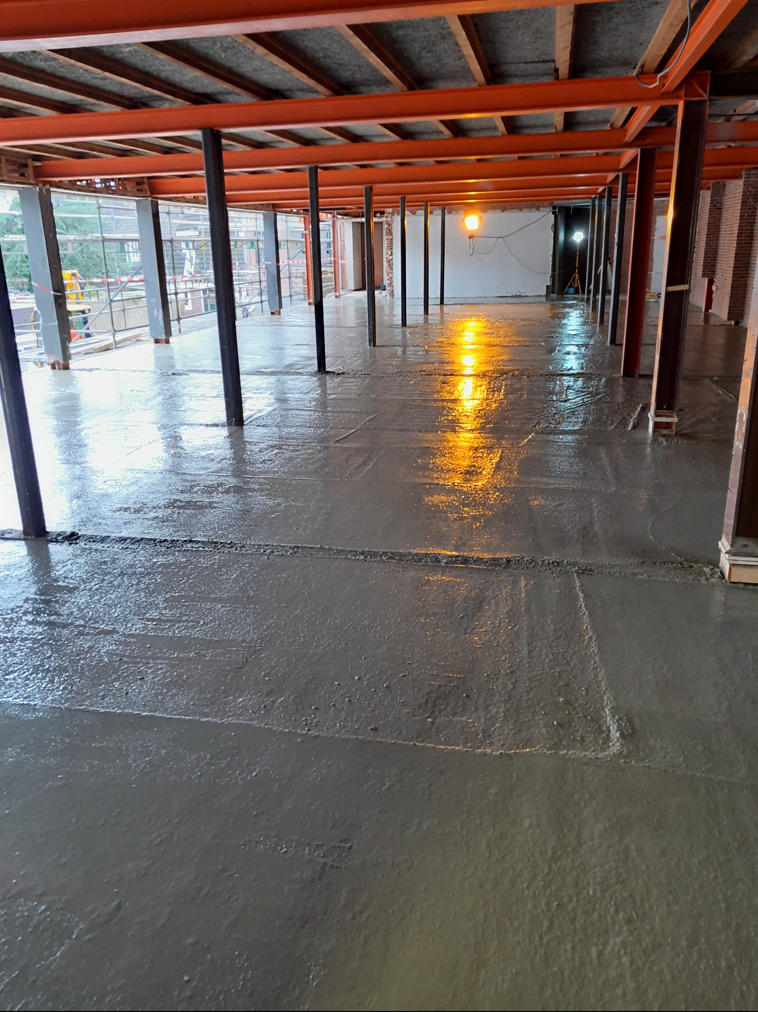 Świeżo wylana, mokra posadzka betonowa w surowym wnętrzu z metalową konstrukcją stropu i kolumnami. Widoczne refleksy światła na gładkiej powierzchni betonu.