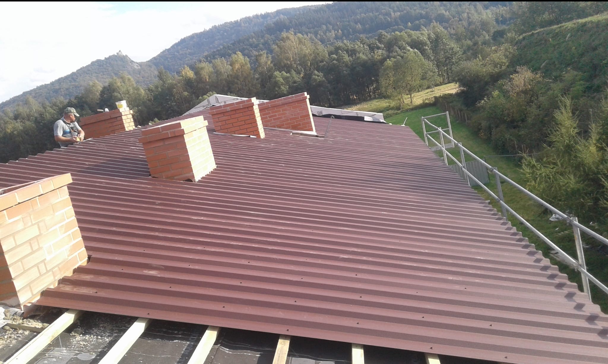 Montaż blachy trapezowej na dachu z kominami w Jeleniej Górze. Widoczny pracownik i rusztowanie. Tło stanowią drzewa i góry.