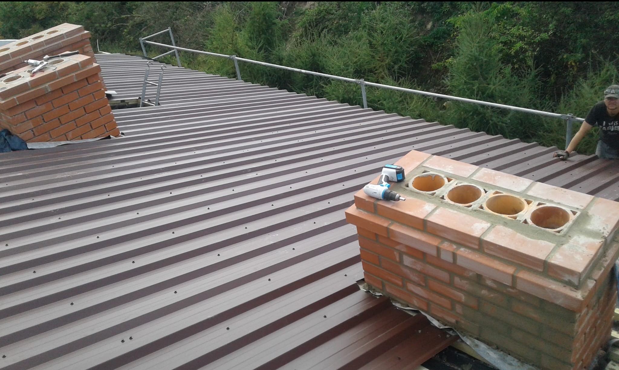 Montaż komina z cegły na brązowym dachu z blachy trapezowej. Widoczny wkrętak akumulatorowy i fragment rusztowania. Praca w toku, zieleń w tle.