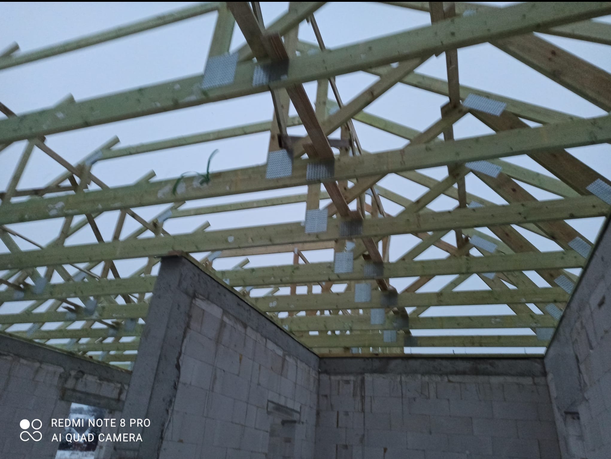 Konstrukcja drewniana dachu z metalowymi łącznikami, widoczna od wewnątrz surowego budynku z białych bloczków. Szare niebo w tle.