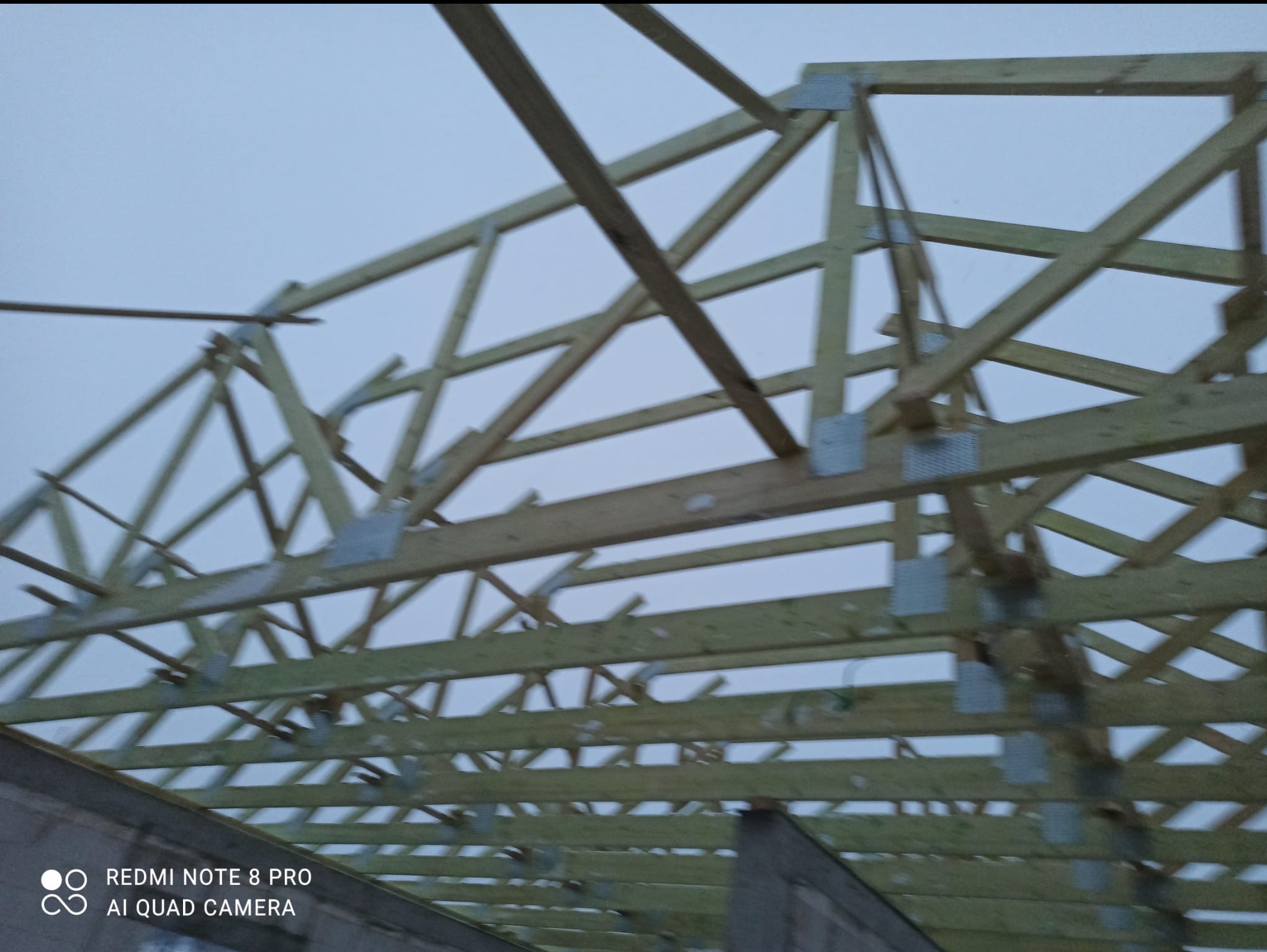 Drewniana konstrukcja dachu z metalowymi łącznikami, widok od dołu na tle pochmurnego nieba. Elementy konstrukcyjne w jasnym odcieniu drewna.