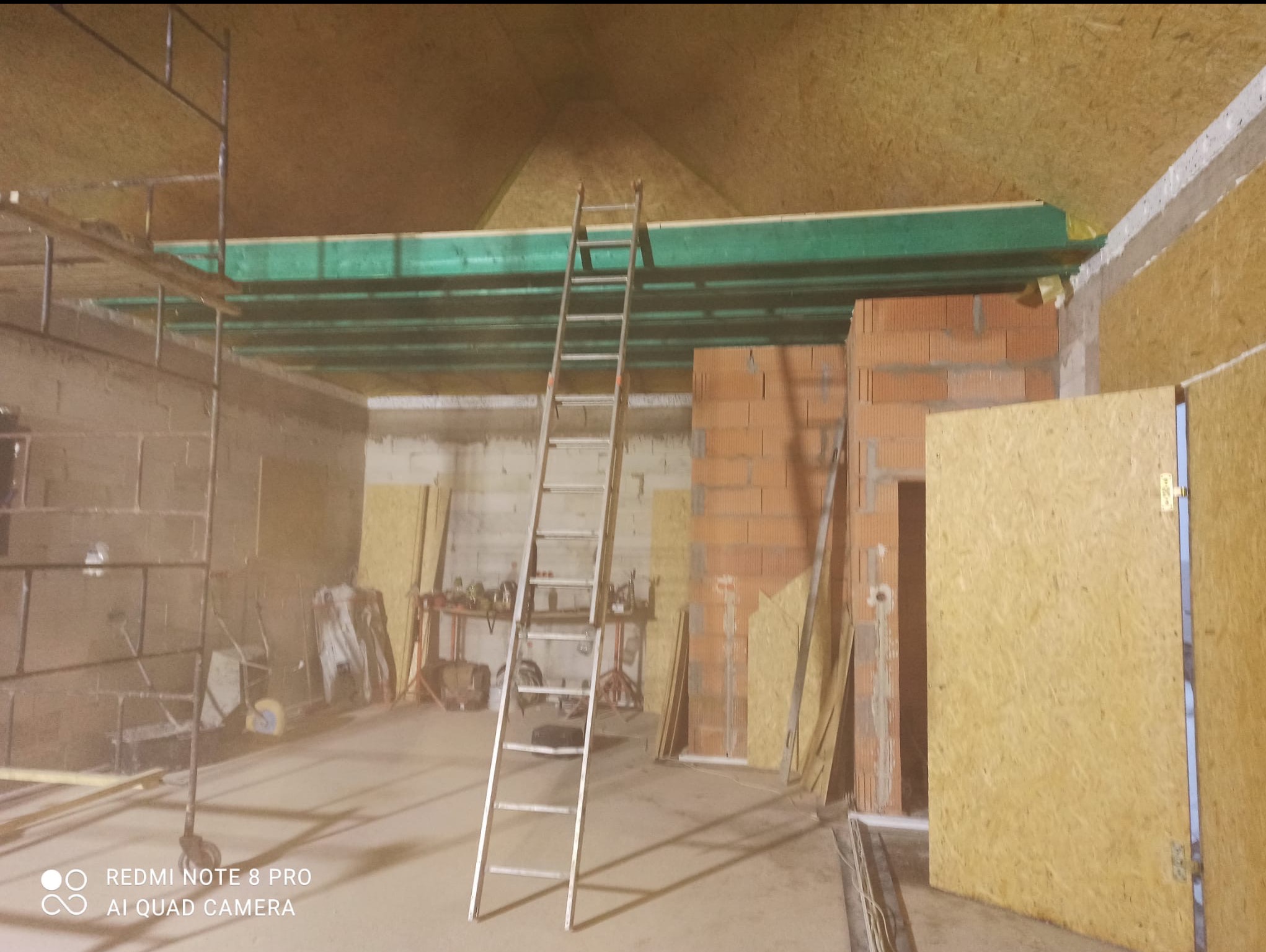 Wnętrze w trakcie prac wykończeniowych: rusztowanie, drabina oparta o ścianę z cegieł, widoczne płyty OSB na ścianach i suficie. Etap budowy antresoli.