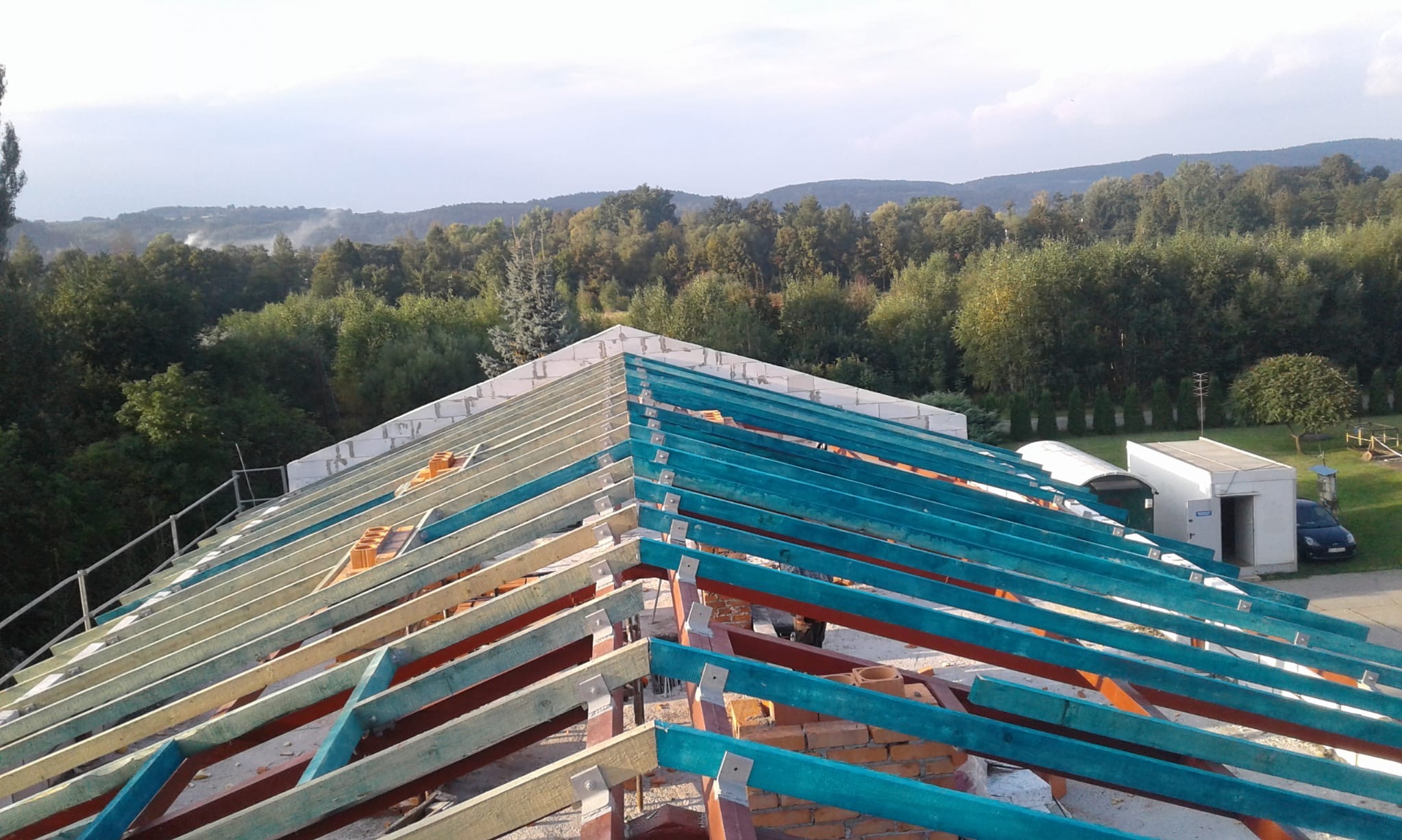Wiązary dachowe w trakcie montażu, widoczne elementy konstrukcyjne i cegły. W tle zalesione wzgórza i budynek gospodarczy.