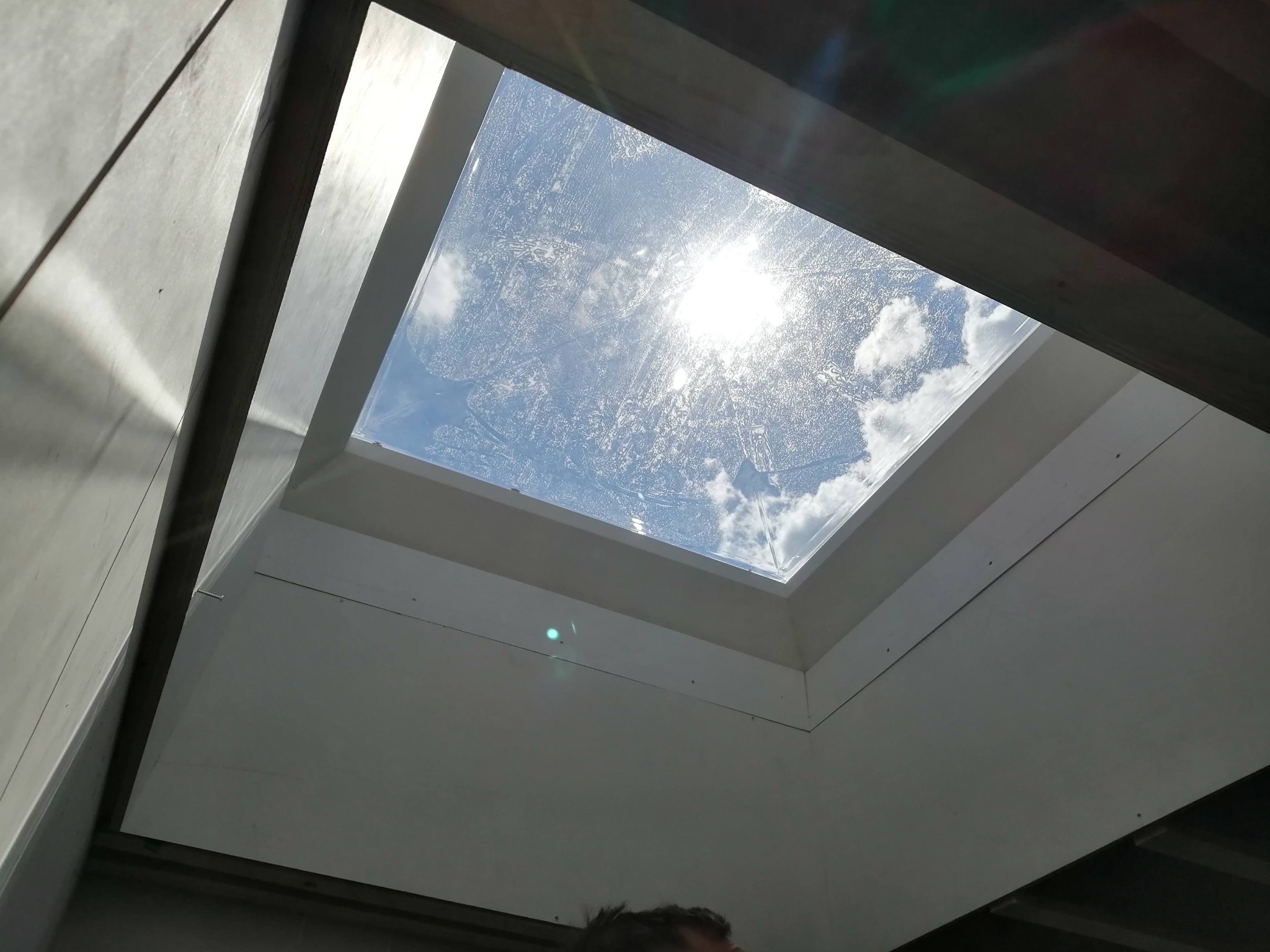 Wnętrze z oknem dachowym, widok na błękitne niebo z chmurami. Białe ściany i rama okna, widoczne ślady montażu. Ujęcie z dołu, fragment głowy osoby w kadrze.