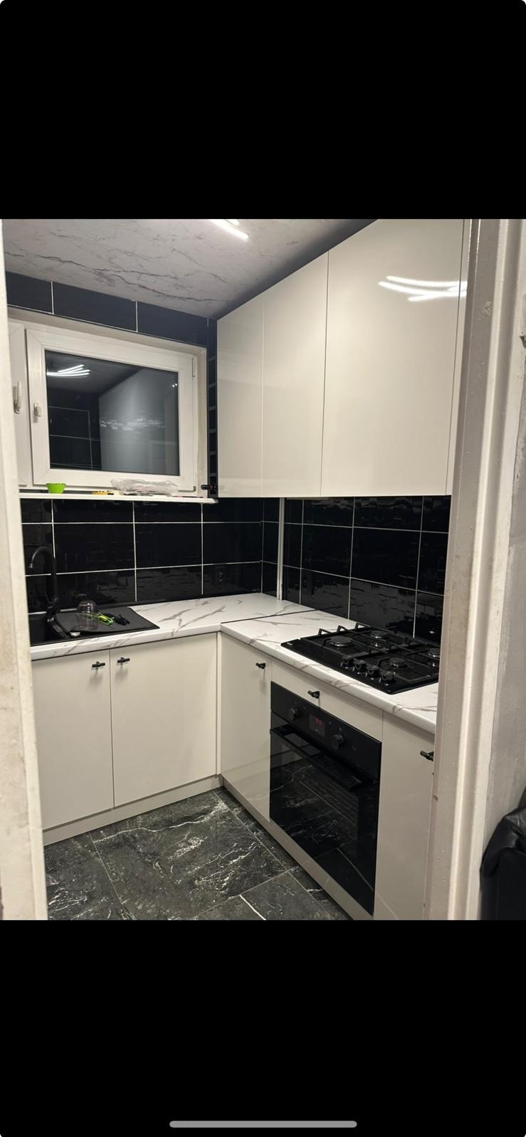 Kompaktowa kuchnia z białymi, lakierowanymi frontami i czarnymi płytkami. Blat imitujący marmur. Małe okno nad zlewem. Nowoczesny design w ograniczonej przestrzeni.