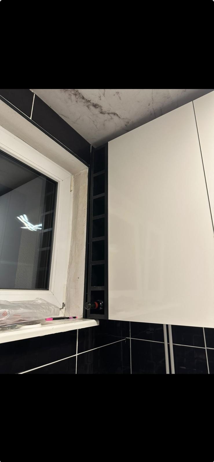 Białe szafki kuchenne z czarnym regałem na wino obok okna. Czarne płytki na ścianie i marmurowy sufit. Butelka wina w regale.