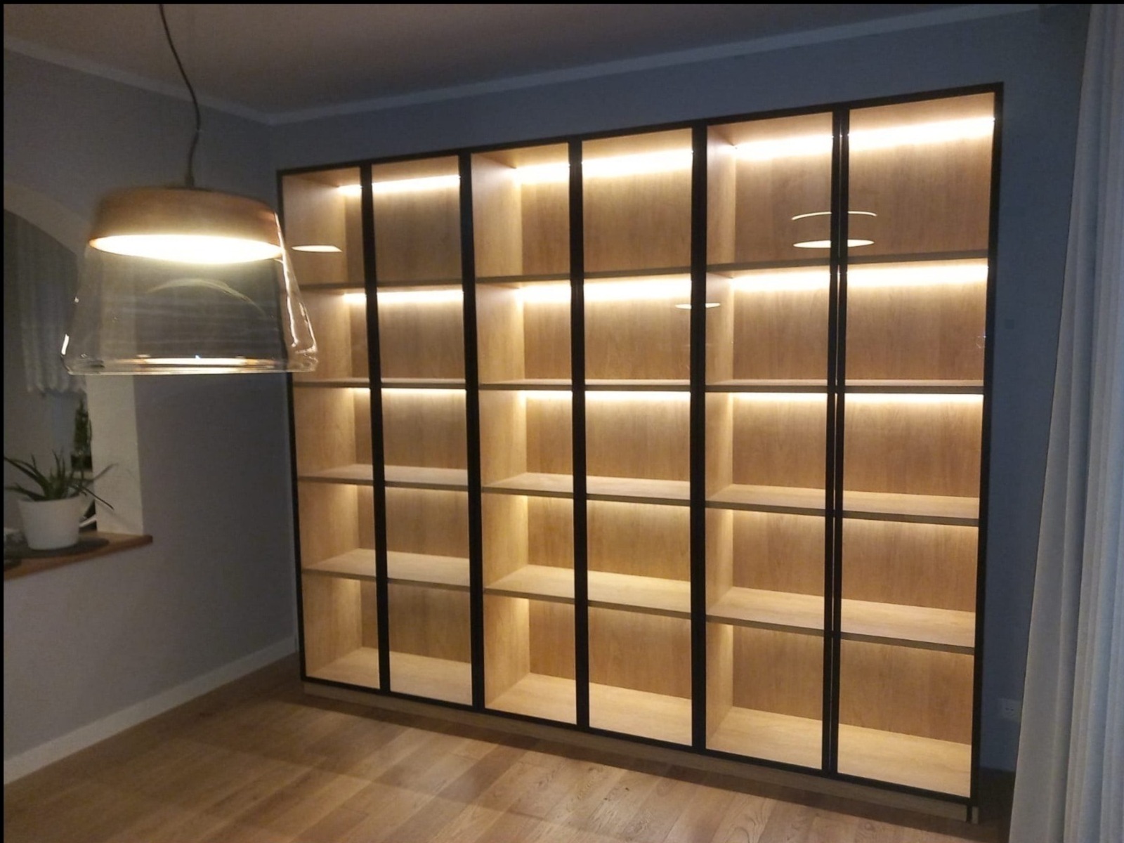 Duża, podświetlana regałowa szafa z jasnego drewna i czarnymi ramami, widoczna w salonie. Oświetlenie LED podkreśla minimalistyczny design i ciepłą kolorystykę wnętrza.