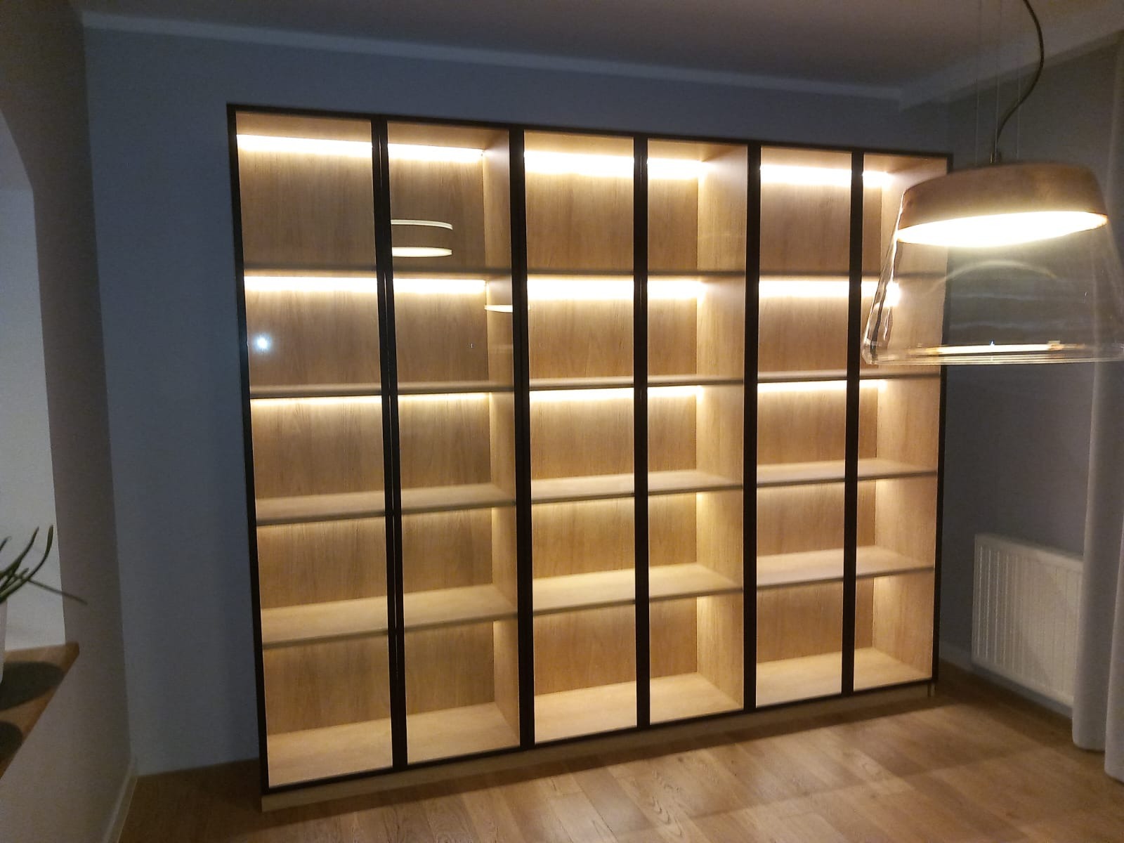 Duża, podświetlana szafa z jasnego drewna i czarnymi ramami, z widocznym fragmentem lampy wiszącej. Minimalistyczny design, ciepłe oświetlenie wnętrza.