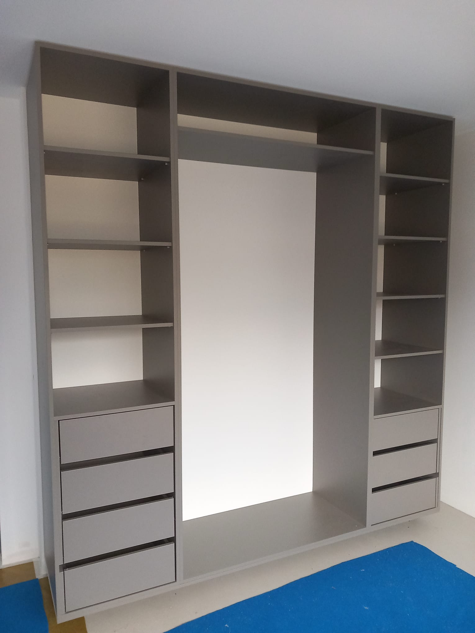 Szara, modułowa szafa wnękowa z szufladami i półkami, widoczna z perspektywy. Minimalistyczny design, świeżo po montażu, z zabezpieczeniem podłogi.