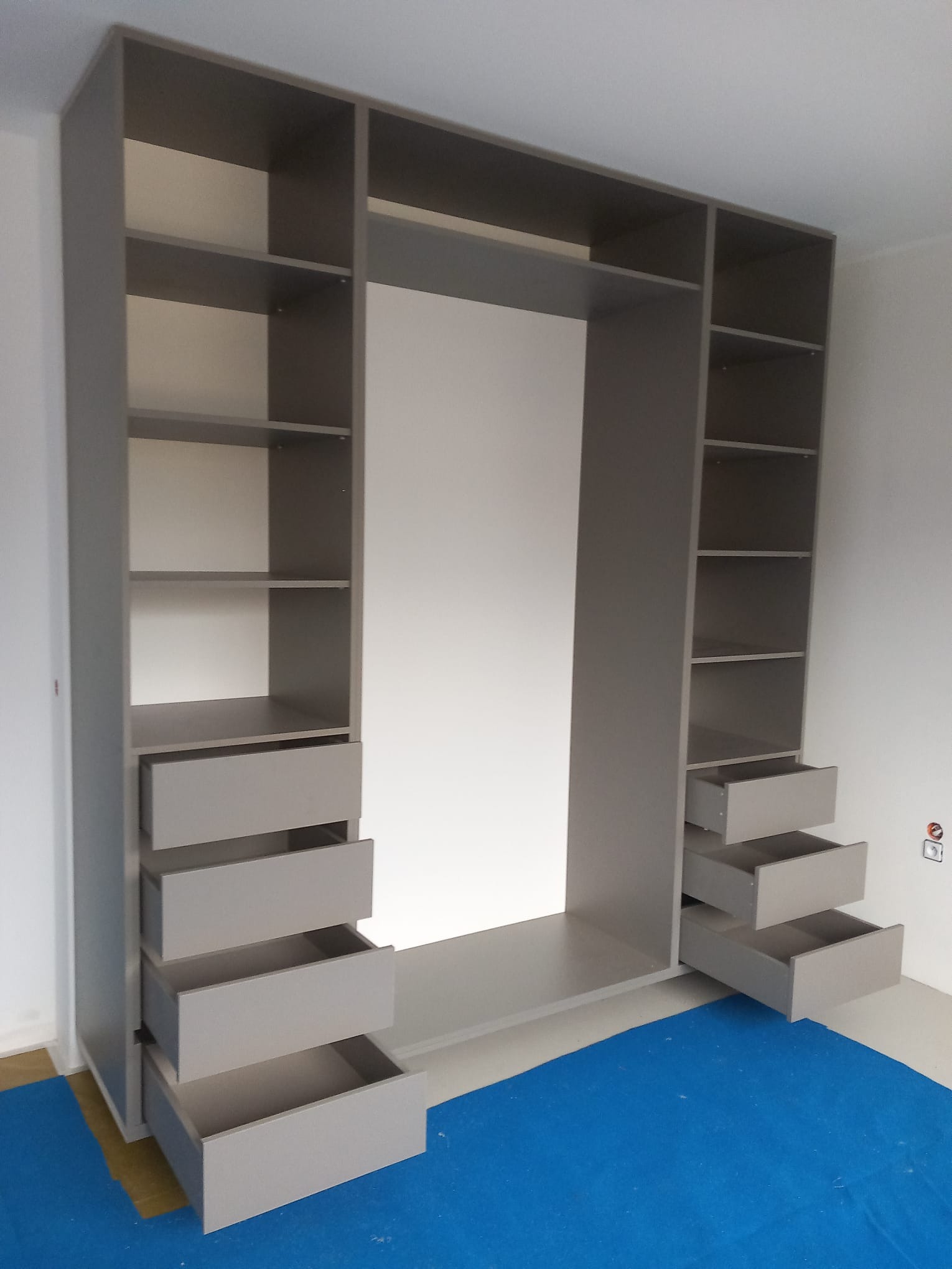 Szara, otwarta szafa wnękowa z półkami i wysuwanymi szufladami, widok z przodu. Na podłodze niebieska folia ochronna, ściany białe. Ujęcie z bliska, wnętrze szafy białe.