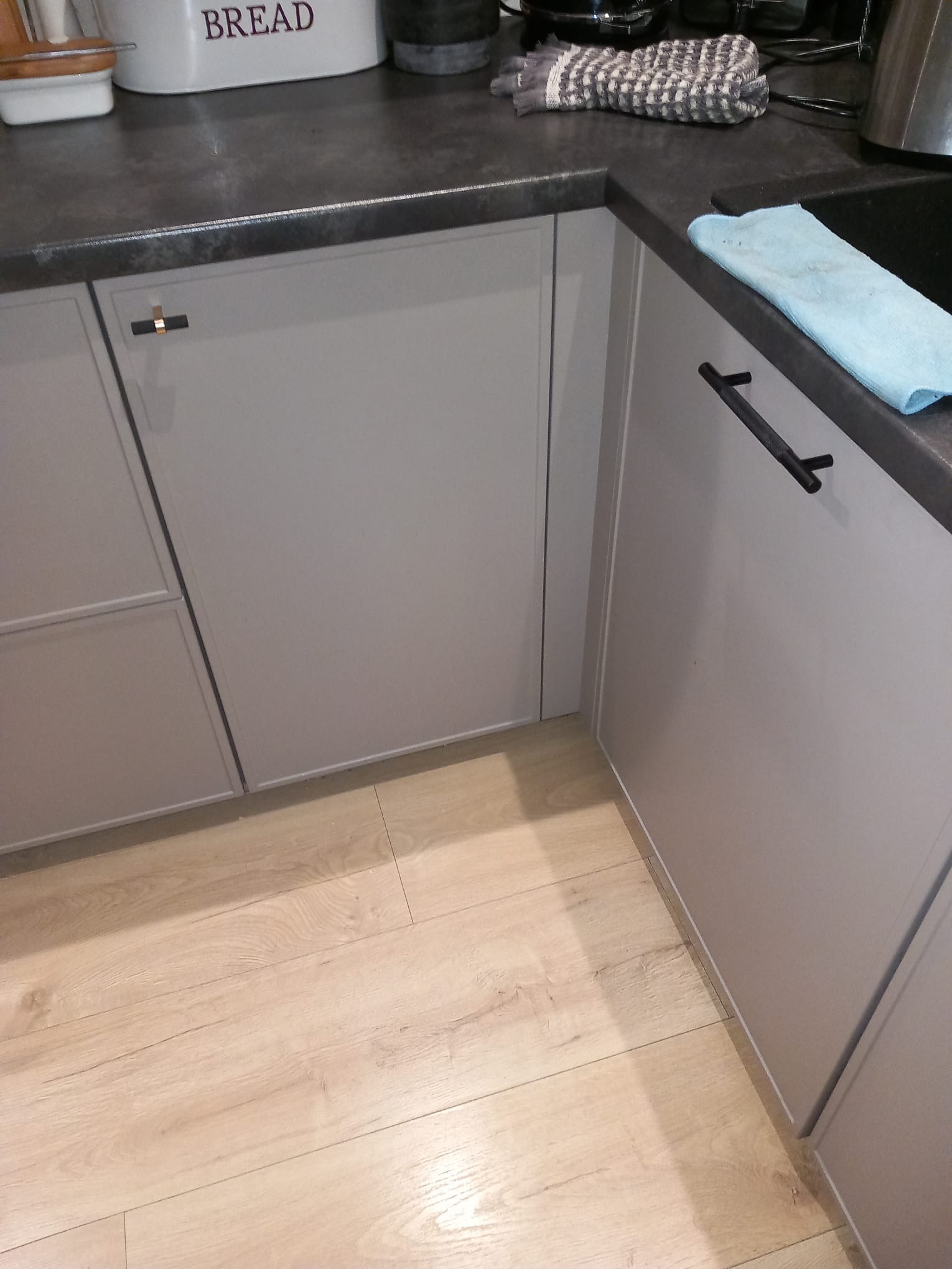 Szare szafki kuchenne z ciemnym blatem i drewnianą podłogą. Na blacie leżą ściereczki i pojemnik na chleb. Widoczne proste, czarne uchwyty.