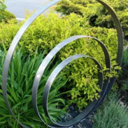 Metalowa konstrukcja spiralna, wkomponowana w zieleń ogrodu. Element dekoracyjny z płaskich, srebrnych prętów na tle krzewów i kamieni. Unikalny detal architektoniczny.