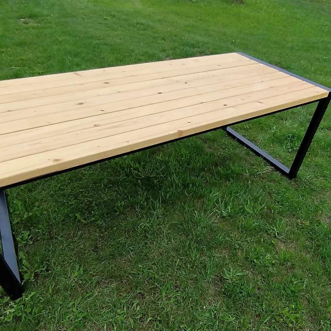 Stół z drewnianym blatem i czarną, metalową ramą o geometrycznym kształcie, ustawiony na trawie. Widoczne detale spawów i naturalna faktura drewna.