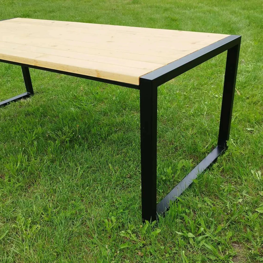Stół z jasnego drewna na czarnych, metalowych nogach, ustawiony na trawie. Minimalistyczny design, widoczne łączenia drewna, spawane elementy konstrukcji.