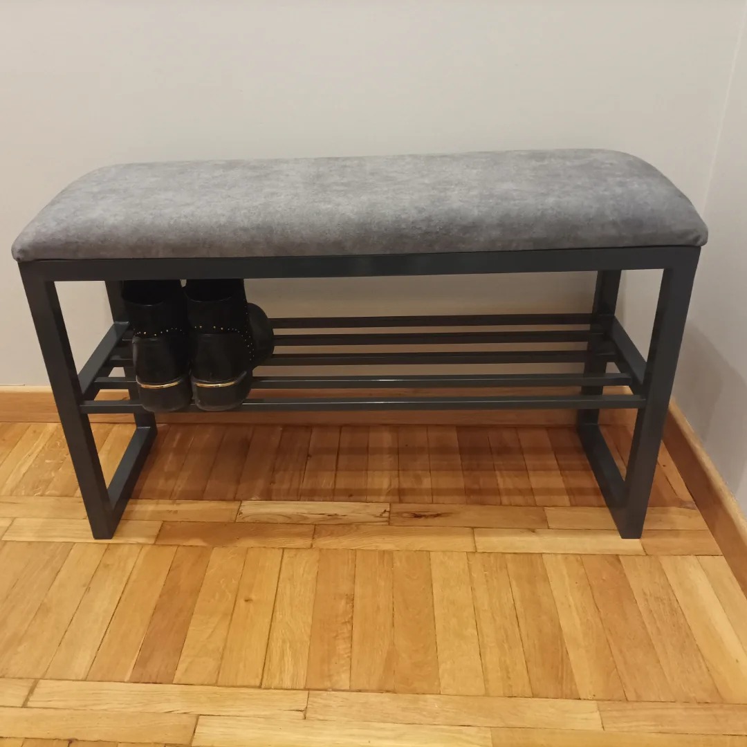 Metalowa ławka z szarą tapicerką i półką na buty, na podłodze z parkietu. Widoczne czarne buty na półce. Minimalistyczny design, wnętrze.