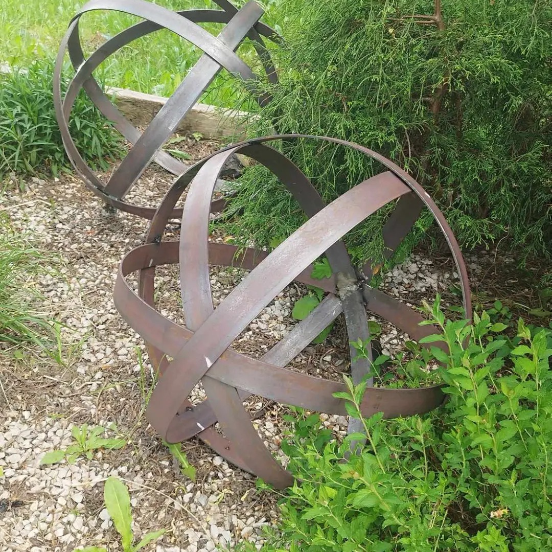 Dwie metalowe kule ogrodowe z płaskowników, z widocznymi spawami, rustykalny styl, otoczone zielenią i kamieniami. Element dekoracyjny w ogrodzie.