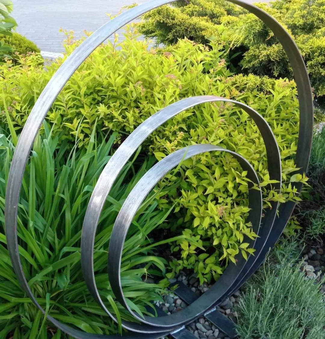 Metalowa konstrukcja spiralna, wkomponowana w zieleń ogrodu. Element dekoracyjny z płaskich, srebrnych prętów na tle krzewów i kamieni. Unikalny detal architektoniczny.