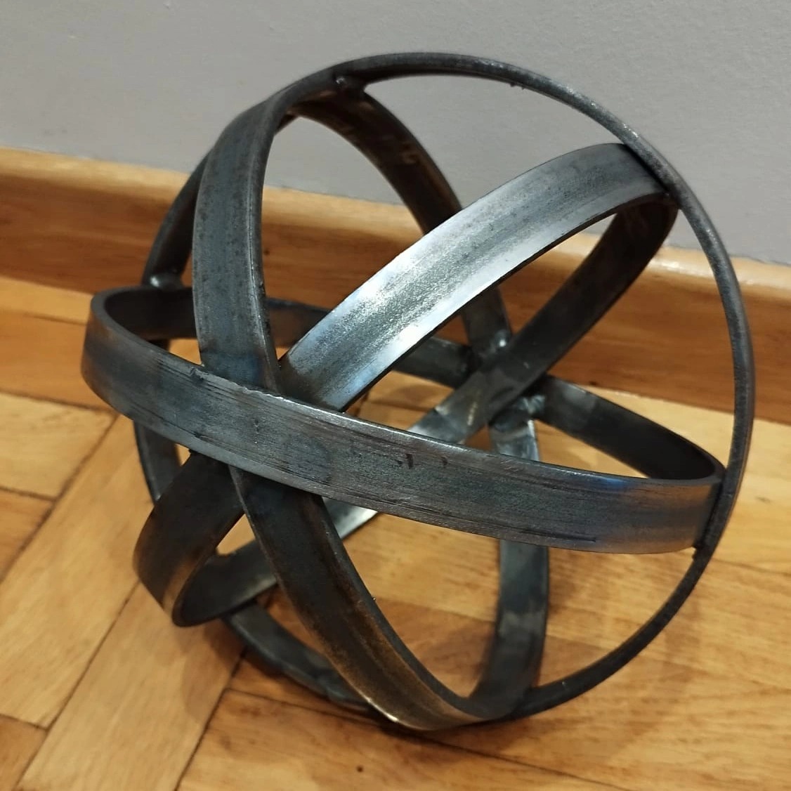 Metalowa kula dekoracyjna z płaskowników, spawana, surowy industrialny styl, widoczne spawy, na drewnianej podłodze, szaro-srebrny odcień.